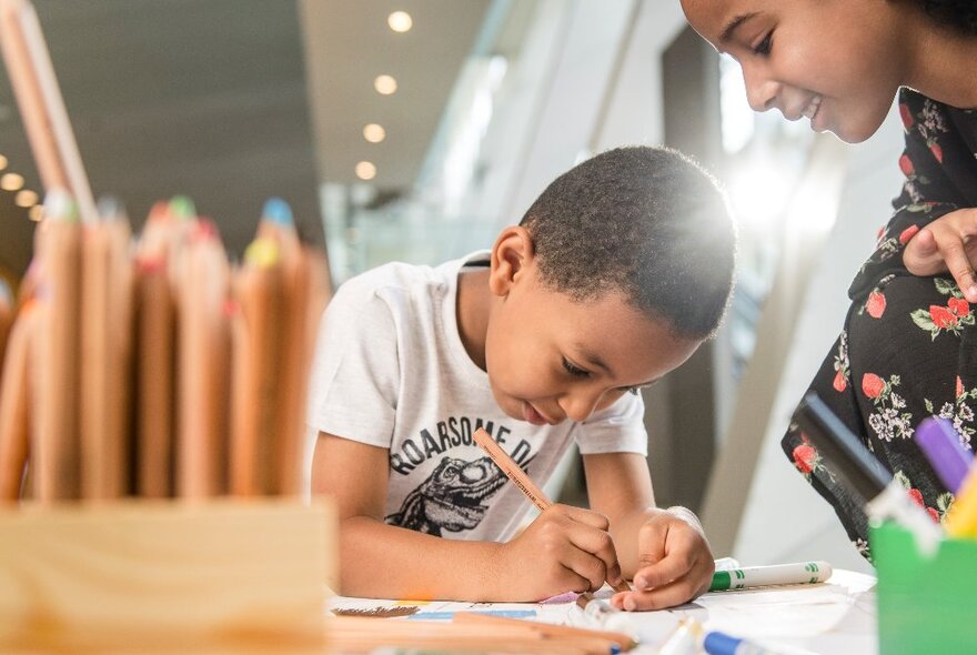 A young boy concentrating on drawing, with a young girl watching and a wooden box of pencils on the table in front of him.