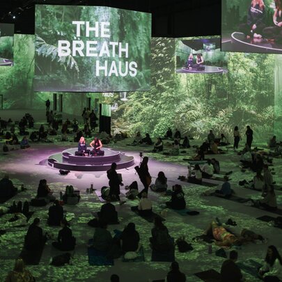 A large green toned area with people gathered for a breathwork class, a projected The Breath Haus sign. 