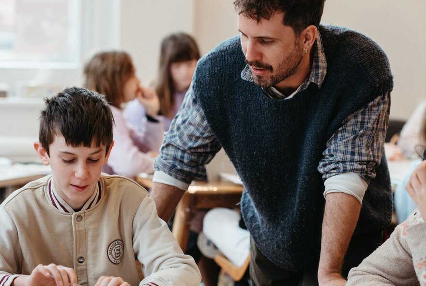 A teacher stands behind seated students who are participating in a creative writing workshop, in a bright and airy studio space.