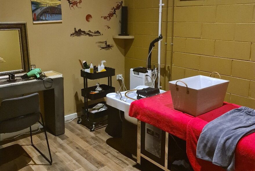 A spa studio room with a long massage table, a head basin at the end of it and other items such as towels, hair dryer and chairs.