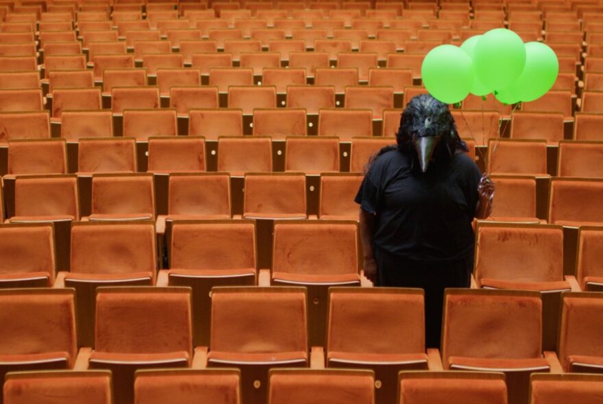 Masked creature holding  green balloons standing amid rows of orange theatre seating.