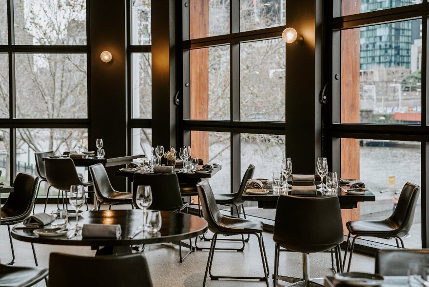 The interior of a restaurant with large floor to ceiling windows overlooking a river, and dining tables set for a meal service.