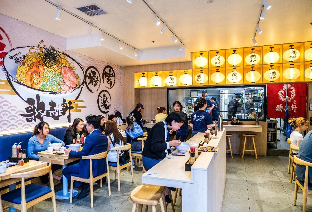 People sitting in a Japanese restaurant with rows of lit lanterns for lighting at the back and an illustration of a noodle bowl on the left wall.
