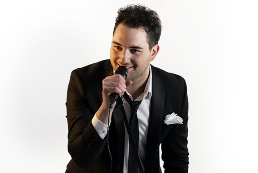 Entertainer Rhys Tolhurst wearing a black suit and tie with a white shirt and pocket square, singing into a microphone; white background.
