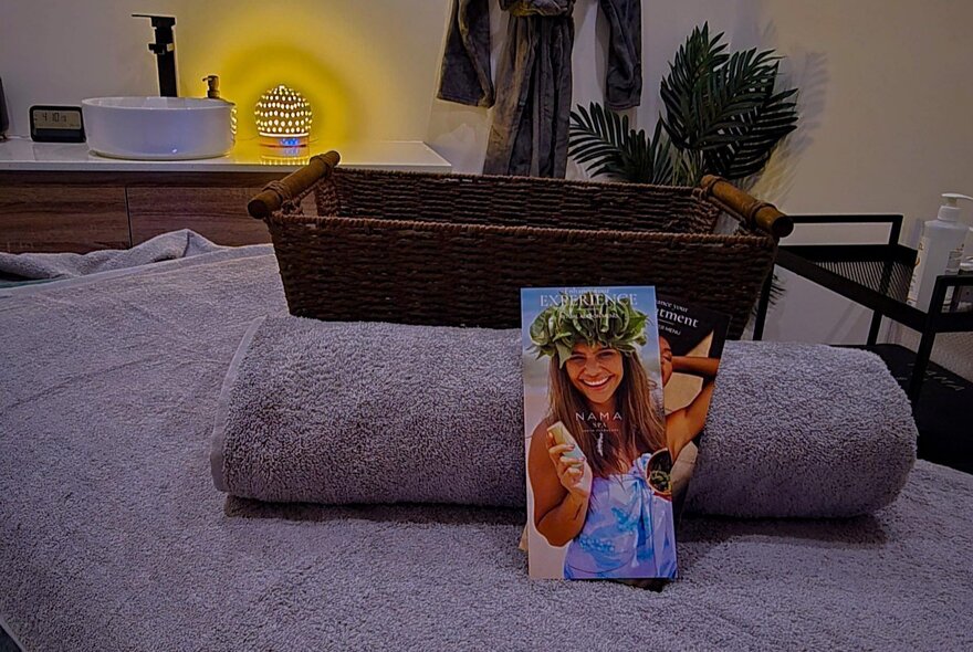 A spa or massage treatment room with a massage bed, rolled grey towel, a brochure and a sink counter in the background.