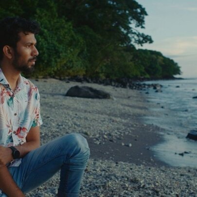 A man in a Hawaiian shirt and jeans sitting on a secluded beach looking out to sea. 