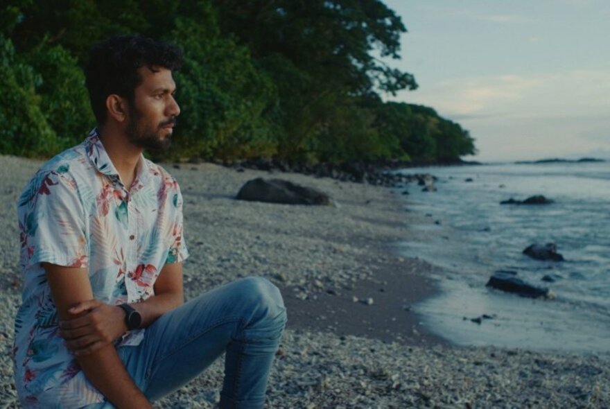 A man in a Hawaiian shirt and jeans sitting on a secluded beach looking out to sea. 