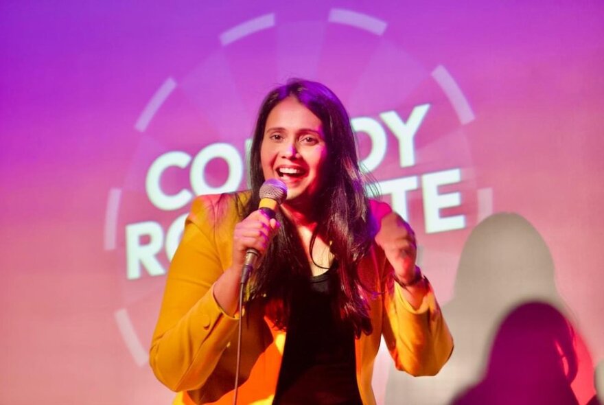 A comedian standing on stage doing stand-up in front of a large screen with lettering and pink tones. 