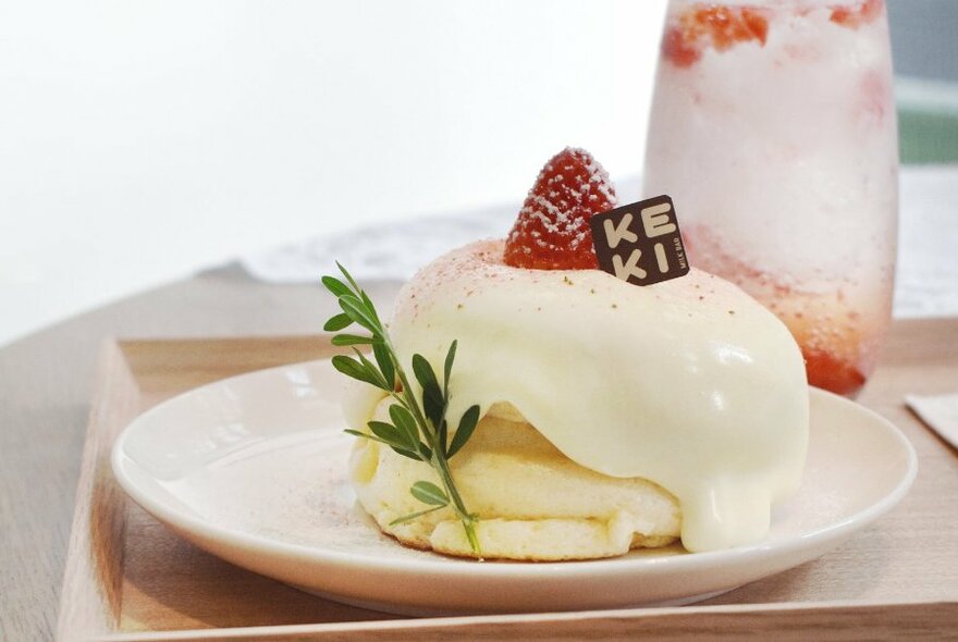 Two soufflé pancakes on a plate covered in custard cream, with a strawberry garnish, and a glass of iced soda in the background.