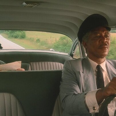 A still image from the movie Driving Miss Daisy, with Morgan Freeman at the wheel of a car and Jessica Tandy in the back seat reading a newspaper. 