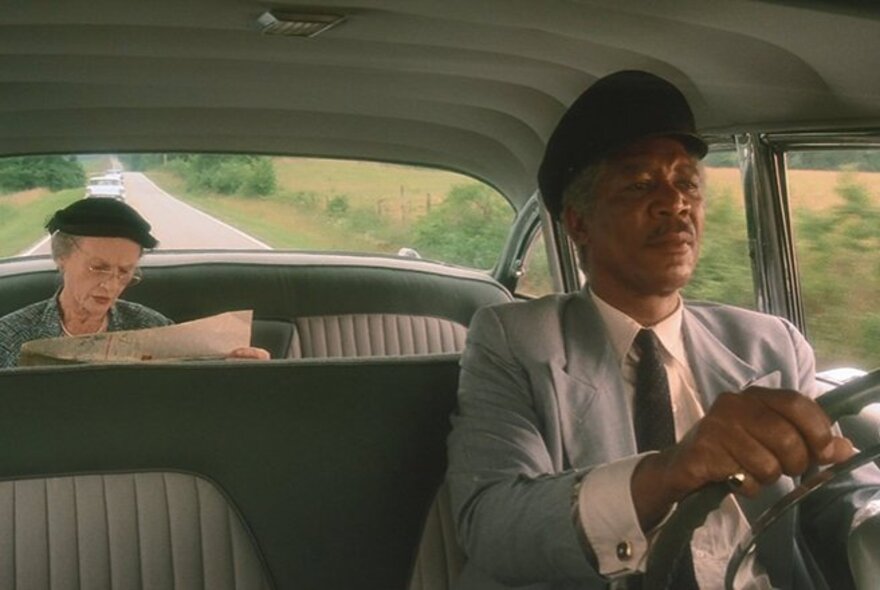 A still image from the movie Driving Miss Daisy, with Morgan Freeman at the wheel of a car and Jessica Tandy in the back seat reading a newspaper. 