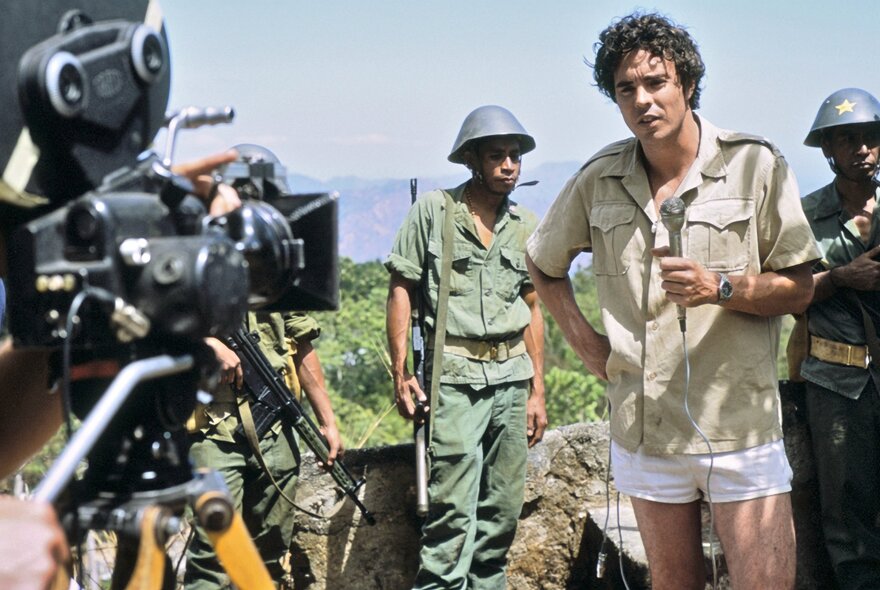 Film still from Balibo, of a reporter being filmed by news cameraman, while armed soldiers look on.