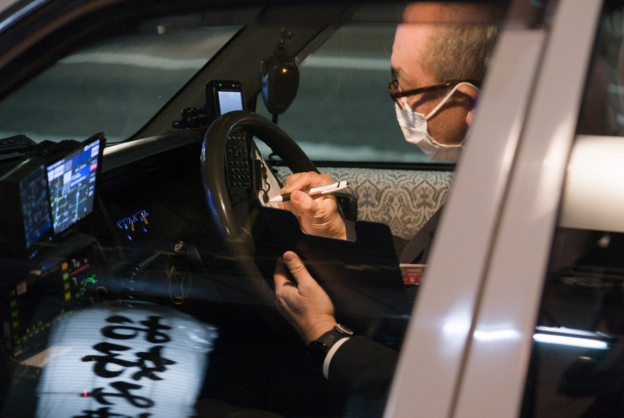 A taxi driver in Japan, wearing a face mask and glasses sits in his car and writes with a pen.