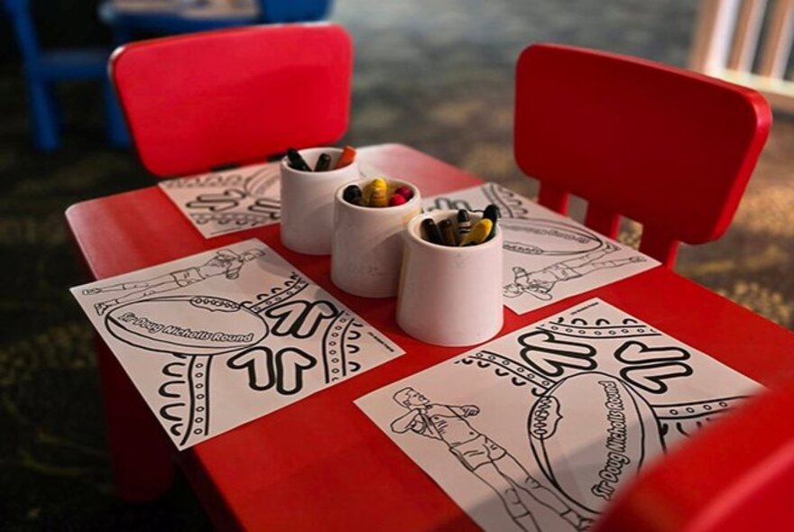 A small red table with two red chairs, with colouring-in stencil sheets and crayons, laid out for a child's craft activity.