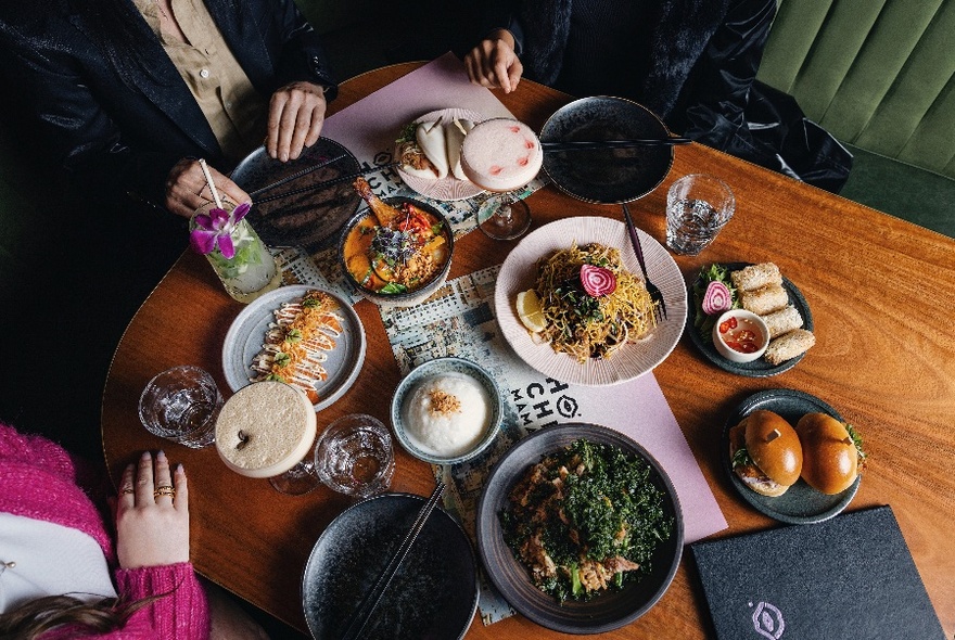 Three people seated around a wooden dining table with several plates of food and drinks resting on large paper menus; view from above.