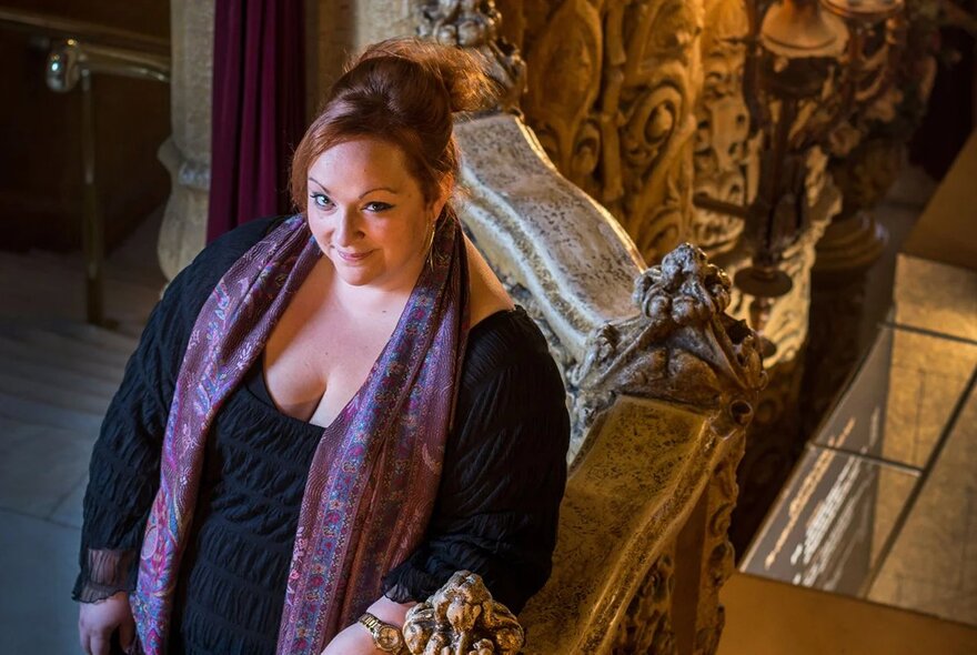 The soprano Helen Dix wearing a black dress with purple trim, her arm resting on a marble balustrade, her eyes raised and looking up.