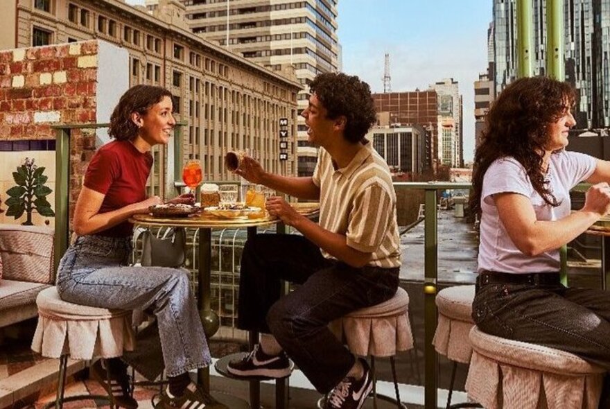 Diners at tables on an outdoor rooftop overlooking Myer and other city buildings, including central couple with drinks and snacks.