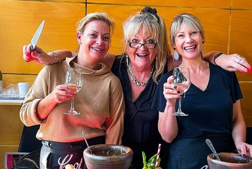 Three smiling women at a Thai cooking workshop with wine glasses, and one holding a chef's knife.