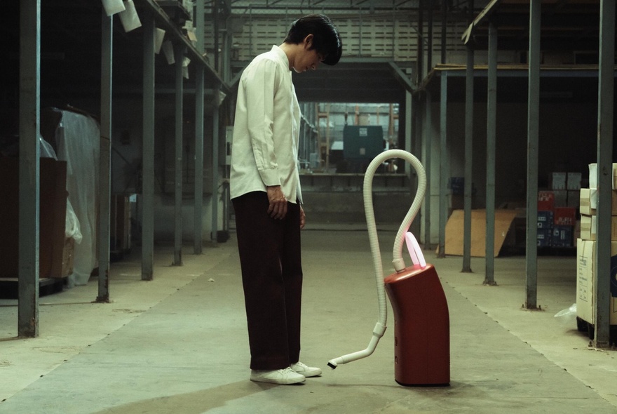 A still image from the film A Useful Ghost, showing a man in a corridor looking down at a vacuum cleaner. 