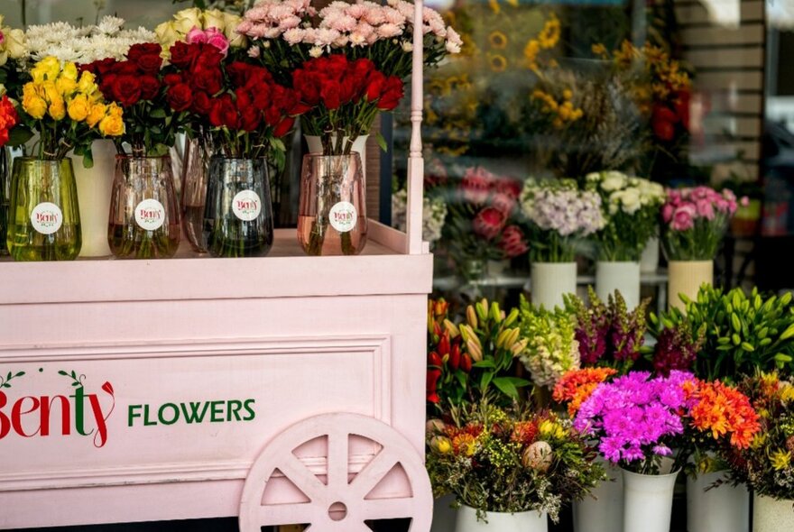 A flower cart and bunches of flowers in white vases outside the front of a florist shop. 