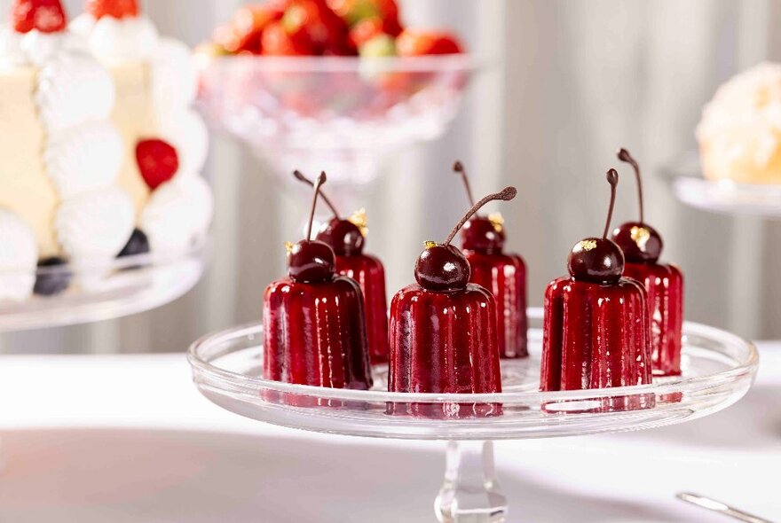 Small red desserts garnished with a cherry, on a cake plate, as part of a dessert buffet in a restaurant.