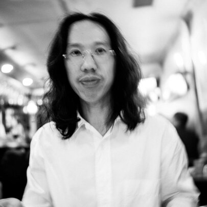 DJ Derrick Gee wearing a white shirt, and glasses, he has shoulder length dark hair and is in a room with a blurred background.