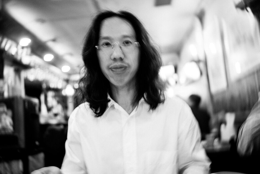 DJ Derrick Gee wearing a white shirt, and glasses, he has shoulder length dark hair and is in a room with a blurred background.