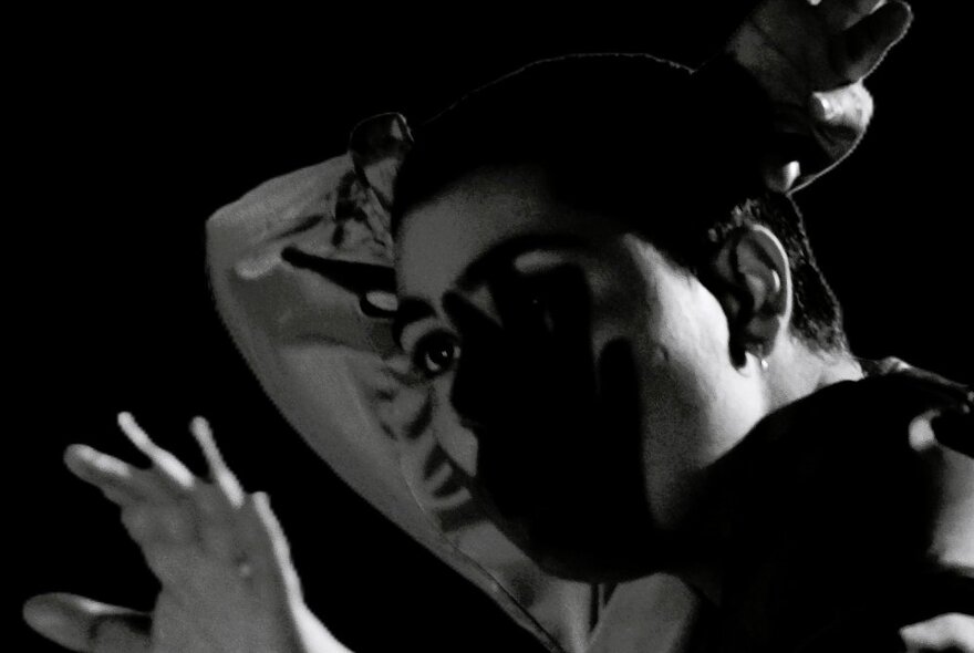 Black and white still from a music video of a person in the shadows, one arm behind their head and the other hand raised in front of their face.