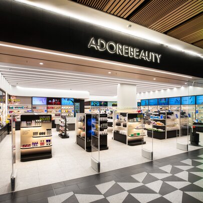 Adore Beauty retail storefront inside a  shopping centre with a large black overhead sign with ADOREBEAUTY printed in white capital letters, and a brightly lit interior with multiple white and black cosmetic display stands and shelves lined with beauty products.