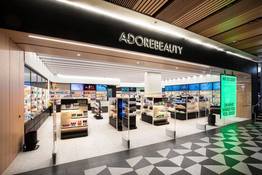 Adore Beauty retail storefront inside a  shopping centre with a large black overhead sign with ADOREBEAUTY printed in white capital letters, and a brightly lit interior with multiple white and black cosmetic display stands and shelves lined with beauty products.
