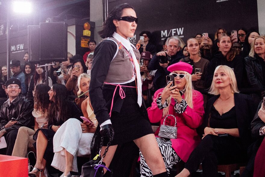 A street-style runway presentation during Melbourne Fashion Week featuring a model in a layered outfit with a grey vest, black skirt, and dark sunglasses, and a seated audience watching.