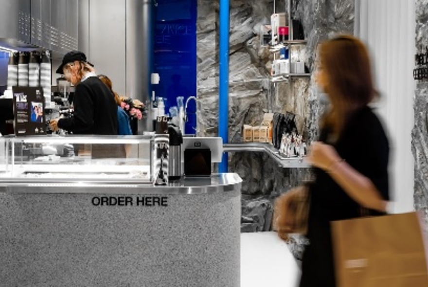 A blurred motion image of a person wearing a cap entering a clean, industrial looking coffee shop with one barista in the background. 