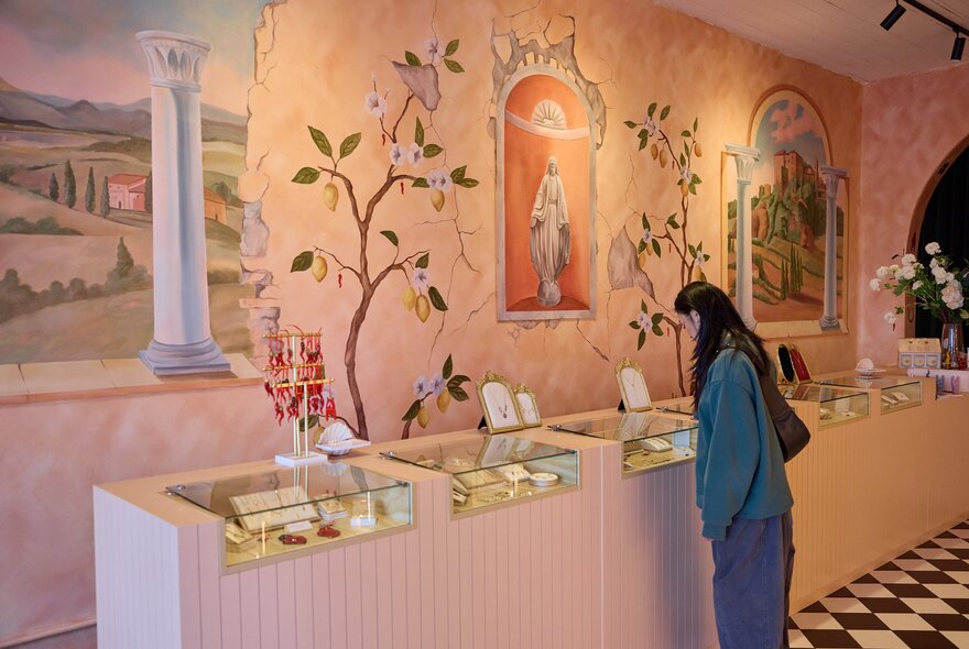 A woman is browsing jewellery at an Italian jewellery shop. The walls are painting pink with Italian frescos on them.