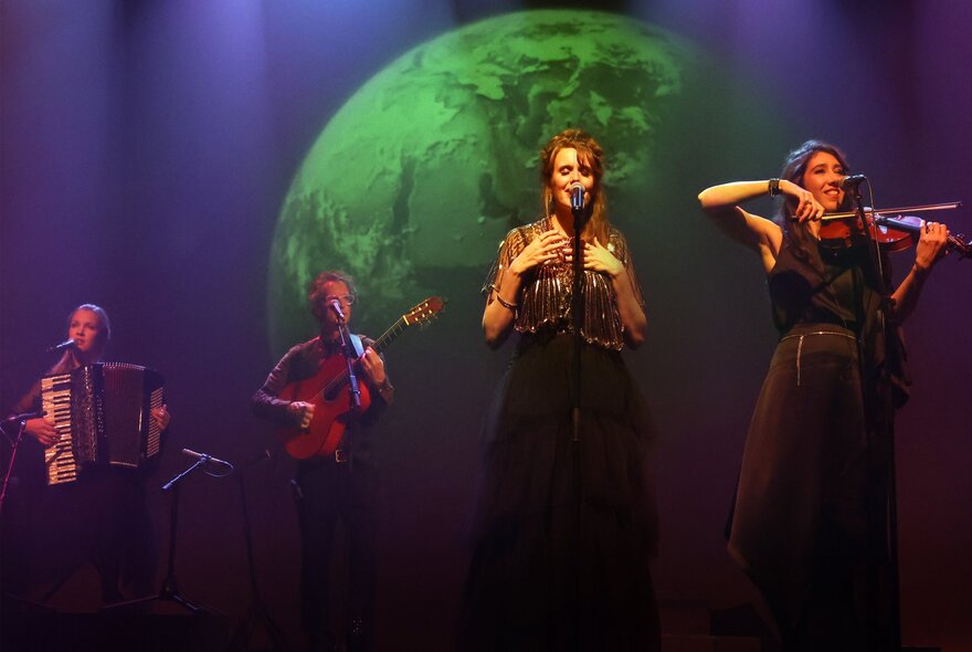 A folk band performing on stage with a woman on fiddle and someone playing a piano accordion, with a green planet featured on their backdrop. 