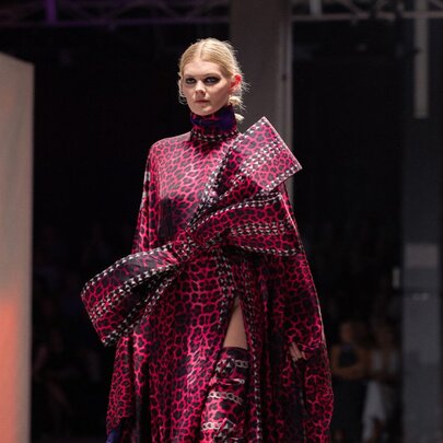 A fashion model wearing a magenta patterned high-neck gown featuring an oversized structured front bow accent on a runway.