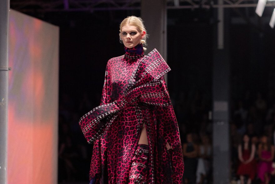 A fashion model wearing a magenta patterned high-neck gown featuring an oversized structured front bow accent on a runway.