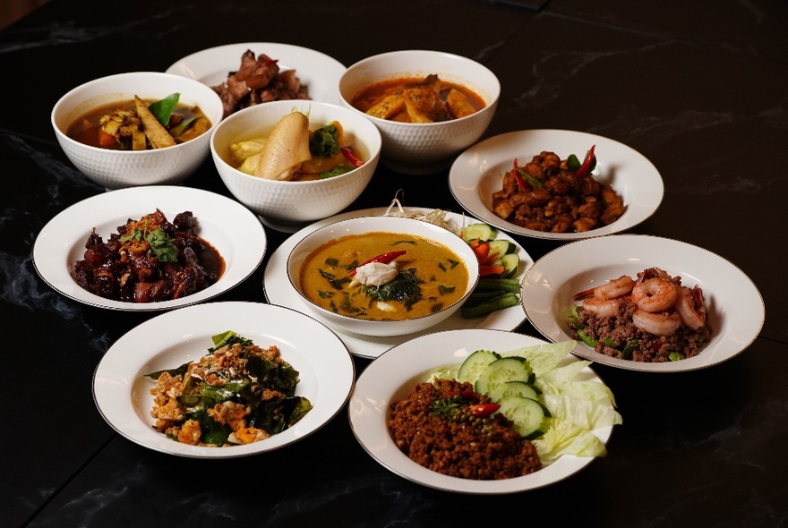 Many shallow white bowls of southern Thai cuisine, including curries, salads and noodles,  displayed on a dark table.