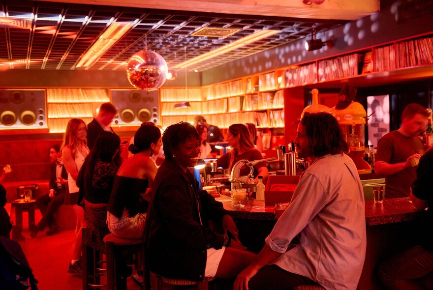 Two friends are sitting in a bar smiling and laughing while drinking cocktails there is a disco ball hanging from the ceiling and records are lined up on the shelves.