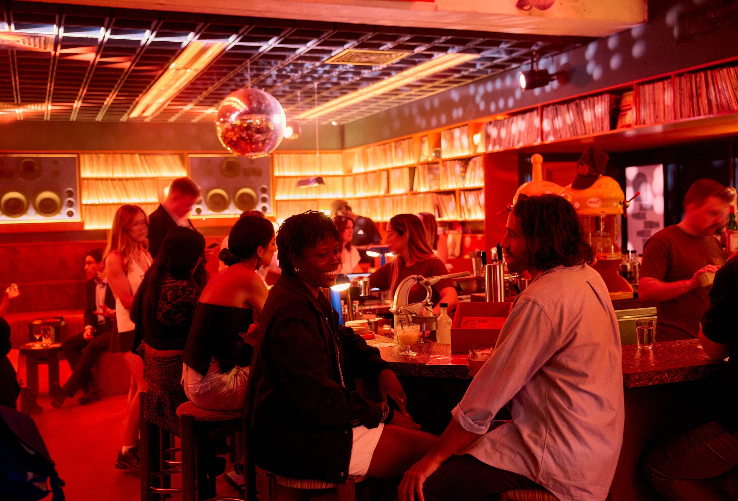 Two friends are sitting in a bar smiling and laughing while drinking cocktails there is a disco ball hanging from the ceiling and records are lined up on the shelves.