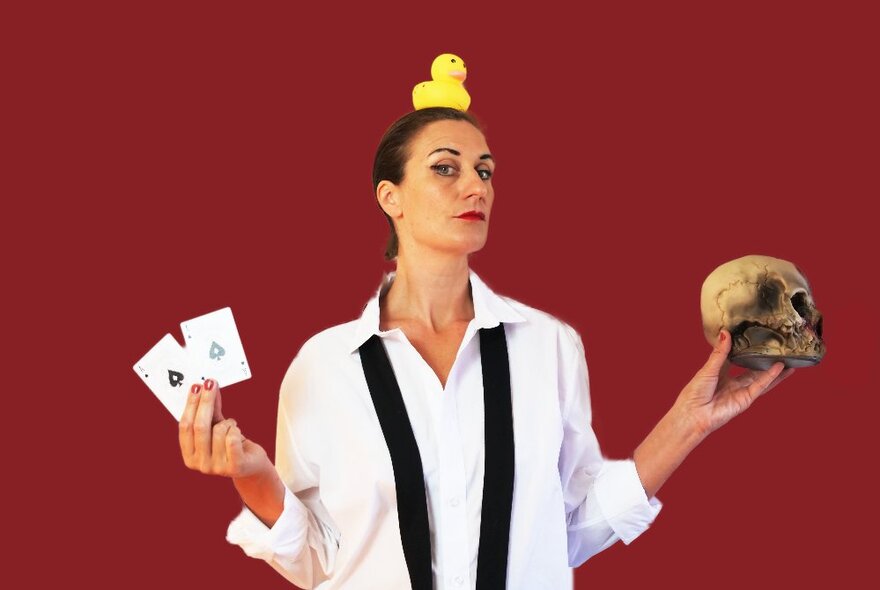 A woman standing with a rubber duck on her head, two aces in one hand and a skull in the other, against a deep red background. 