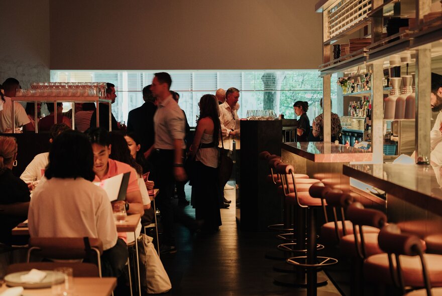 The dimly-lit interior of a restaurant with patrons eating and a line of empty barstools at the bar.