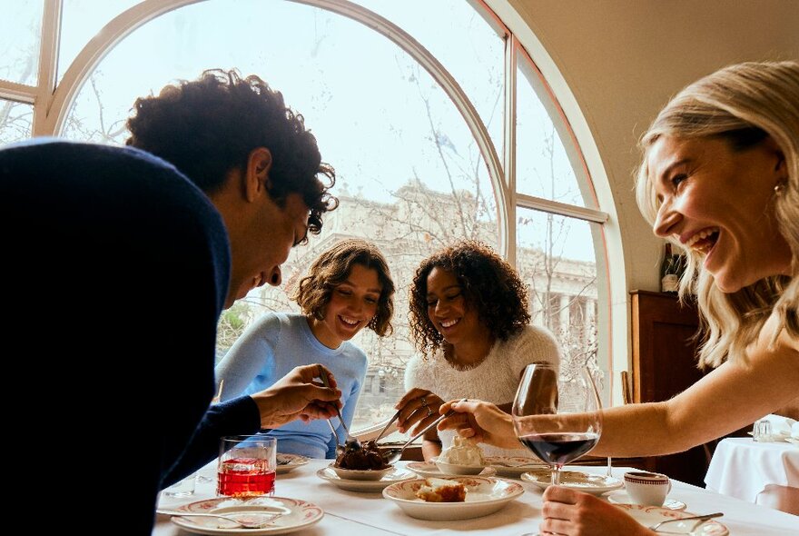 People seated at a table in front of an arched window overlooking Spring St, choosing from dishes of food and drinks.