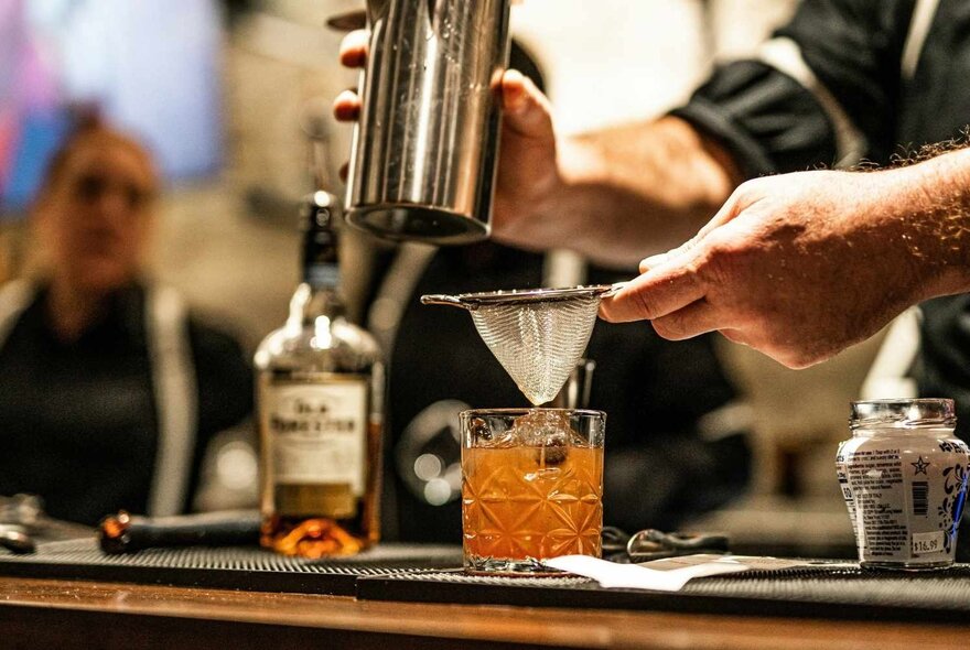 A cocktail being prepared at a bar, featuring a bottle of whiskey and a bartender straining a drink into a glass.