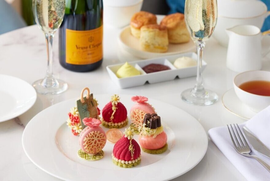 A white plate with six intricately decorated sweet treats on a white linen tablecloth, with two glasses of Champagne, and a china cup of tea and scones in the background.