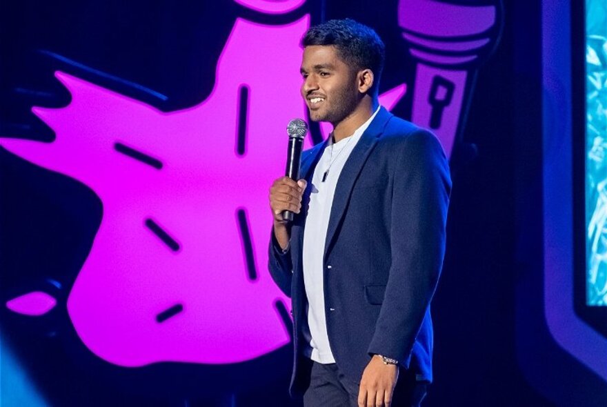 Comedian Nadun Hetti, on stage with a bright purple shape on the wall behind him.