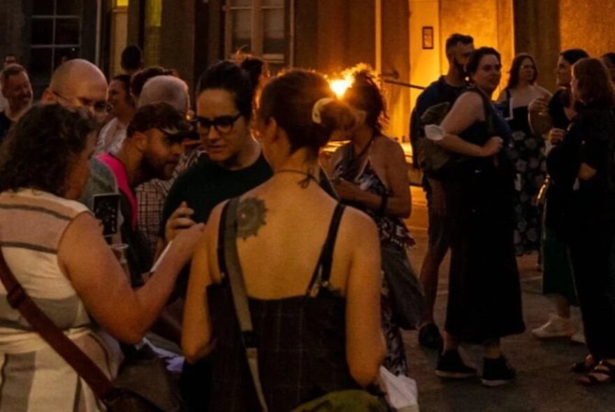 A large group of people mingling in a room lit by laps, the woman closest has a large tattoo on her back. 
