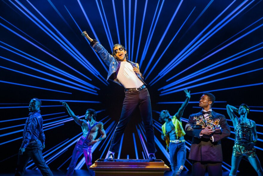 A dancer on stage in a white t-shirt and black pants, in the role of Michael Jackson, with musicians and dancers alongside, in the stage production of MJ the Musical.