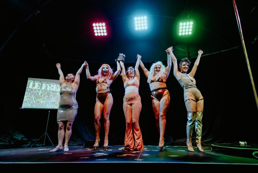 Five performers on a stage, in burlesque-circus style costumes, holding hands and with their arms raised above their heads.