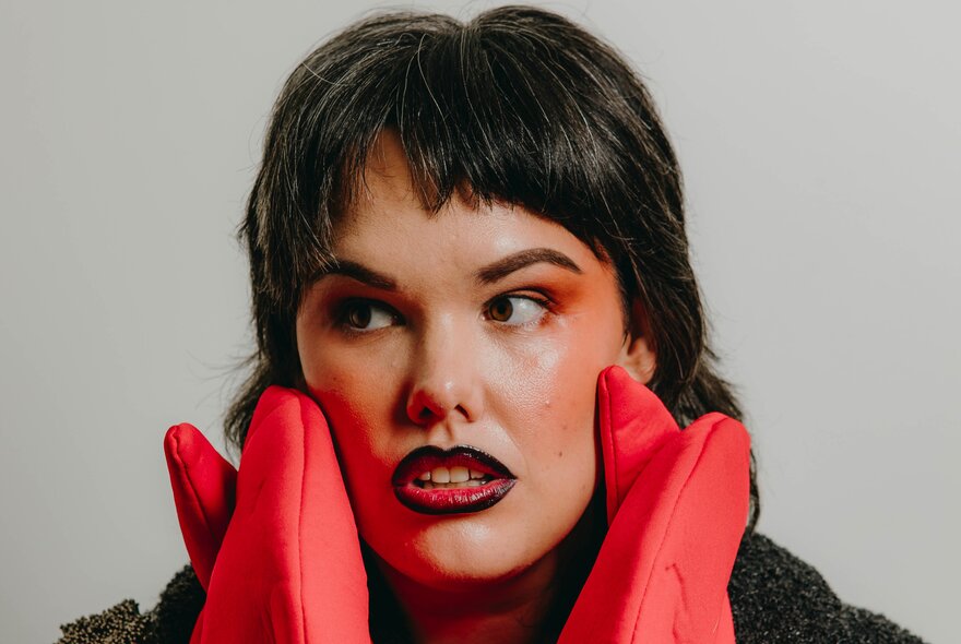 A close-up portrait of a person with dark hair, a short fringe, looking off to the left with an open-mouthed expression, framing their face with both hands while wearing bright red, quilted oven mitts.