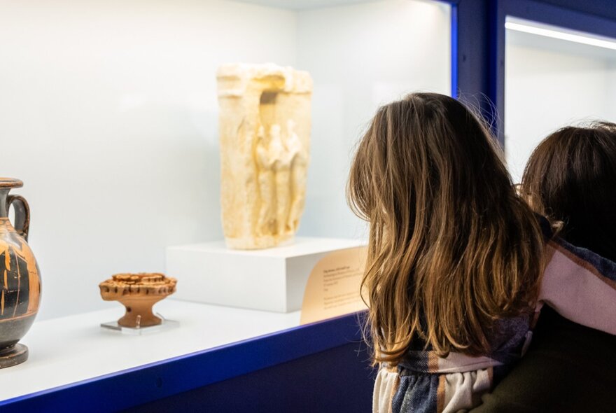 Two children looking at ancient Grecian artefacts on display in a glass cabinet.